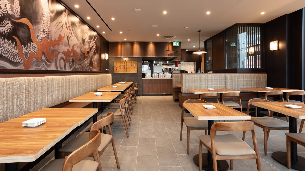 Modern restaurant interior with wooden tables, beige chairs, minimalist decor, warm lighting, and open kitchen concept