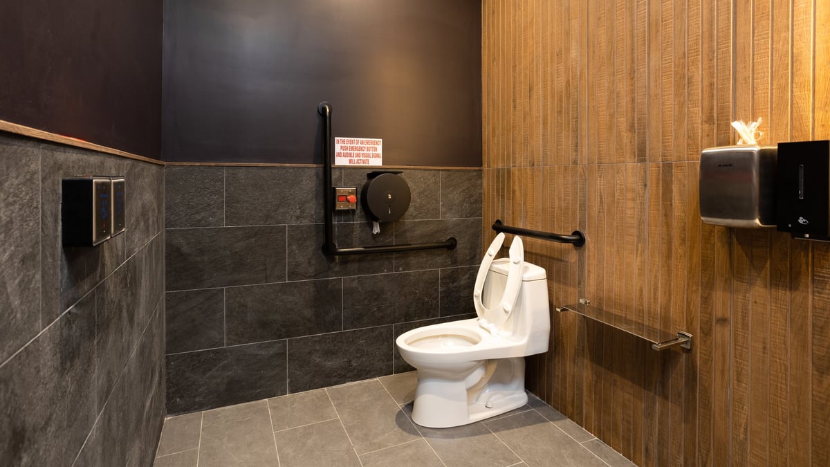 Modern public restroom with dark tile and wood paneling, featuring a white toilet and sink area with grab bars