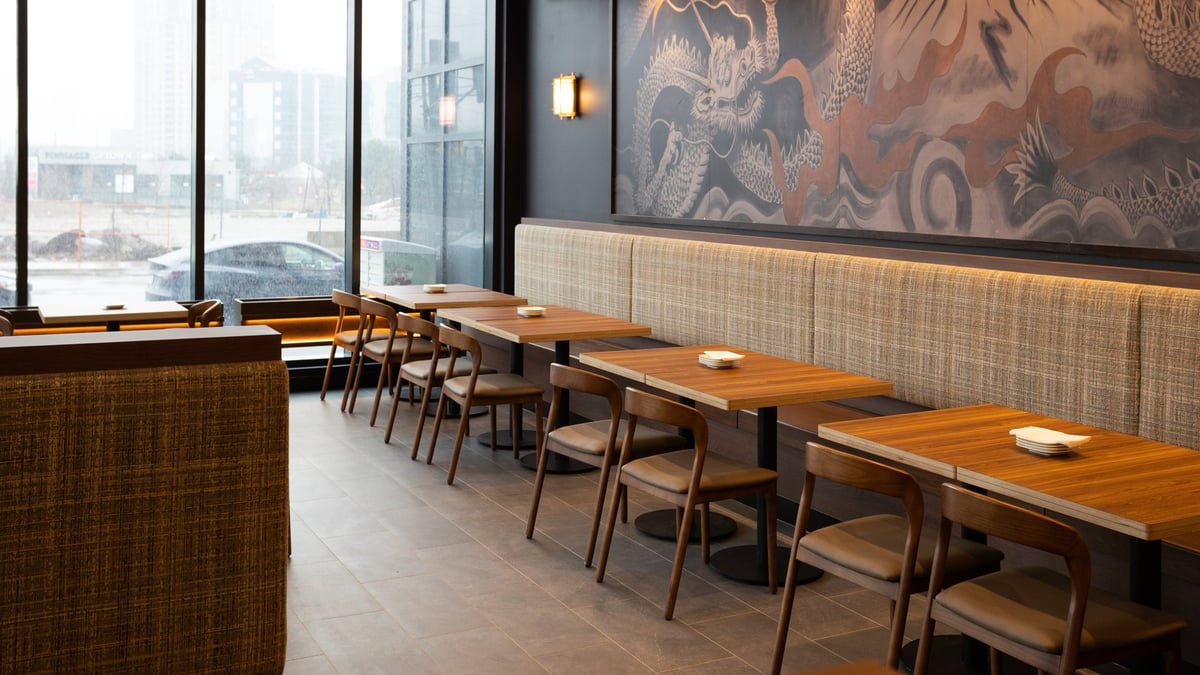 Modern restaurant interior with wooden tables, black chairs, floor-to-ceiling windows overlooking city, and decorative mural on wall